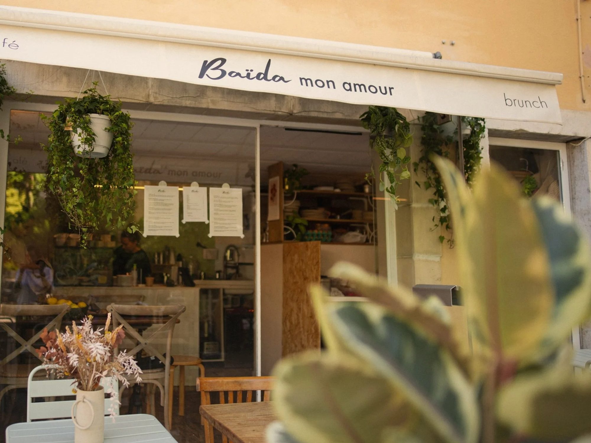 La devanture de Baïda mon amour, façade crème et store sombre, plantes suspendues et tables en terrasse
